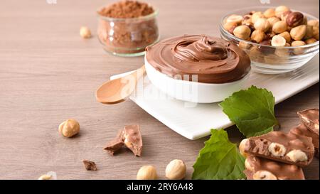 Containers with chocolate cream and hazelnut with nuts, leaves and and chocolate portions on wooden table. Elevated view. Stock Photo