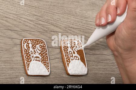 Closeup of decorating Christmas gingerbread by piping bag on wooden background. Female hand painting winter church on sweet baked cookies. Xmas pastry. Stock Photo