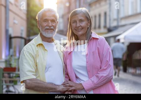 Portrait of an older couple, a man and a woman standing together on a city street, hugging and holding hands, smiling and posing for the camera. Stock Photo