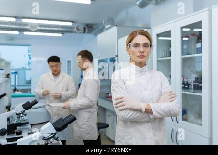 Portrait of a young female scientist standing confidently with her arms crossed on her chest and looking seriously into the camera, behind her male colleagues are discussing research. Stock Photo