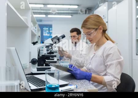 A young female scientist is making research and analysis notes while sitting in a laboratory. Nearby, a colleague works under a microscope. Stock Photo