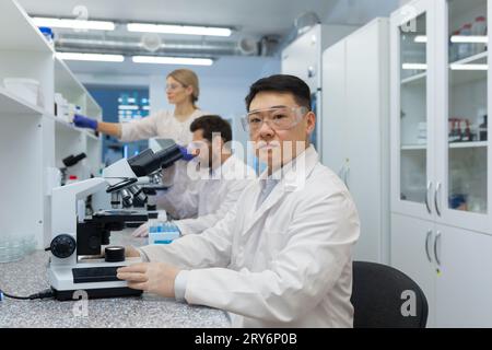 A group of young medical students and scientists are studying in the laboratory of the university. Asian young man sits behind a microscope and looks seriously at the camera. Stock Photo