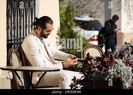 Handsome bearded man in glasses and white suit sitting with smartphone in street cafe Stock Photo