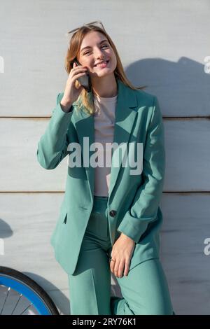 Young empowered ginger-haired woman in green suit talking on smartphone standing at marble background Stock Photo