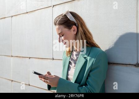 Young empowered ginger-haired woman in green suit browsing on smartphone standing at marble background Stock Photo