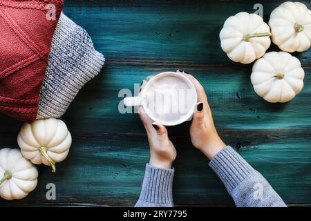 Flatlay of woman's hands holding hot, steaming cup of coffee over a rustic green table with autumn sweaters and white pumpkins. Table top view. Stock Photo