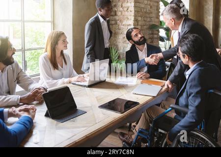 Young bearded man and senior silver-haired individual seal a deal amid diverse colleagues, including an inclusive member in a wheelchair. Bright offic Stock Photo