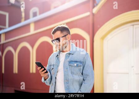 Handsome young adult man in sunglasses and denim jacket looking at mobile phone in his hand while walking street. Stock Photo