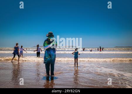 Essaouira, Morocco - August 3, 2023: Woman on the shore at the beach fully covered with typical clothes and hat. City life Stock Photo