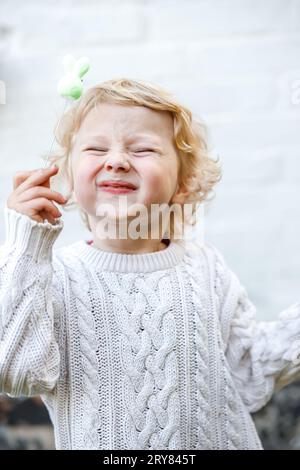 Portrait of a little girl squinting her eyes and holding a toy on a stick. Child in warm knitted sweater on white wall background outdoors. Stock Photo