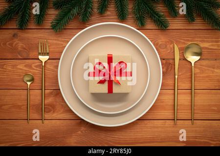 Christmas table setting with festive decorations. New year vintage greeting card. Plate with gift box and golden cutlery on brown wooden board. Top vi Stock Photo