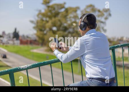 Man in shirt with headphones using his mobile phone leaning on the railing of a pedestrian bridge. Stock Photo