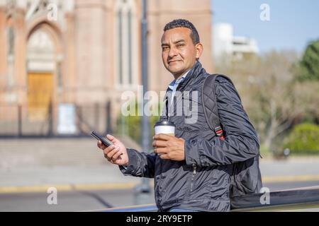 Portrait of Latino man using his mobile phone and holding a disposable cup of coffee in a public park. Stock Photo