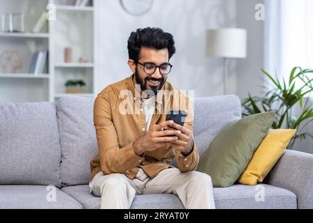 Young hispanic man sitting at home on sofa in living room of house, happy and smiling man holding phone, using smartphone app, typing message and watching video, online shopping. Stock Photo