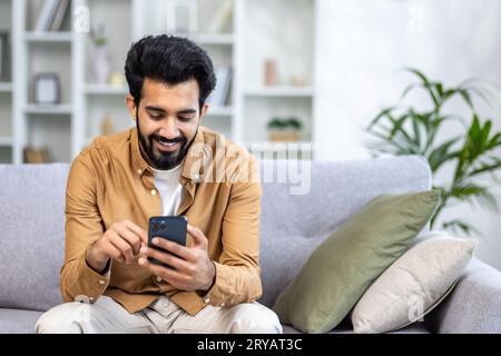 Young hispanic man sitting at home on sofa in living room of house, happy and smiling man holding phone, using smartphone app, typing message and watching video, online shopping. Stock Photo