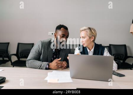 Portrait of focused multiethnic business partners looking at laptop screen, discussing software application. Young black male intern employee Stock Photo