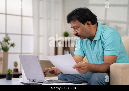 Indian middle aged man checking financial bills, insurence or mortgage documents on laptop while sitting on sofa at home - concept of Financial Stock Photo