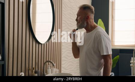 Caucasian retired handsome elderly man senior mature old 60s male bearded pensioner with gray hair looking at mirror reflection combing beard with Stock Photo