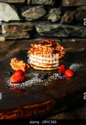 Tasty pancakes with strawberries, syrup, sugar and bacon on the top - darke background. Pancakes with fruits on dark wooden table. Stock Photo