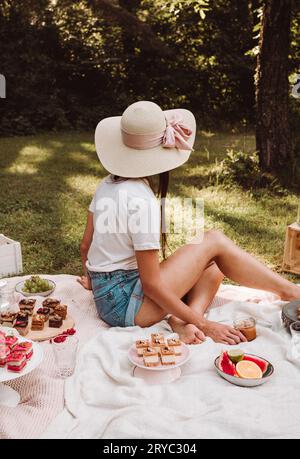 Portrait photo of woman sitting on the blanket with cakes, sweets and other food in nature - picnic style. Lifestyle image - female model with straw h Stock Photo