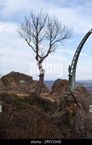 Donnersberg, Germany - April 9, 2021: Tree next to side of Adlerbogen, Eagle Arch, with a statue of a man on a hill with rocks on a cloudy spring day Stock Photo