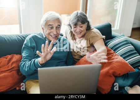 Senior couple with white hair sitting on the sofa, waving towards a laptop. Emphasizing elderly using technology to stay connected with loved ones rem Stock Photo