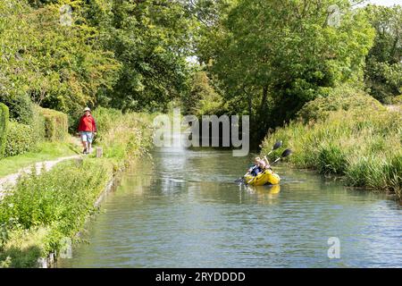 A man walking along a canal path with a man and a woman paddling in a kayak on the Basingstoke Canal near Odiham in August. Hampshire, England Stock Photo