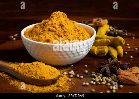 A bowl of yellow turmeric powder surrounded by wooden spoon with turmeric powder, dry spices including star anise, nutmeg, cumin, coriander seeds, dry Stock Photo