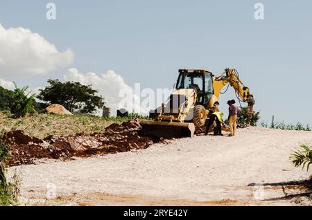 A front-end loader with its wheeles lift from the ground is attended by men working on the road project. Stock Photo