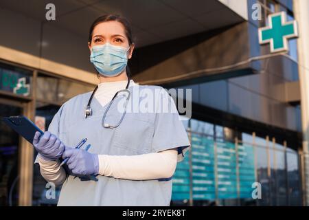 Young female doctor standing in front of healthcare facility, wearing protective face mask and PPE e Stock Photo