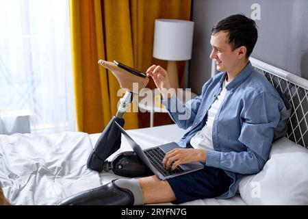 impaired man with artificial leg prosthesis checking messages and working online having remote job, at home in bedroom. excited guy laughing, smartpho Stock Photo