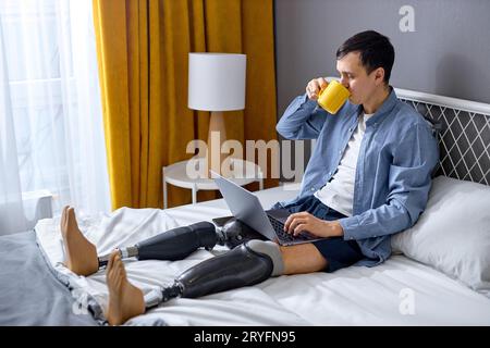 Young worker with disability and special needs sit on bed and work on laptop. Serious and concentrated. Handicapped Caucasian male using laptop for wo Stock Photo