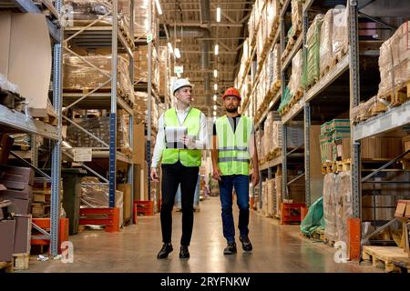 Retail Warehouse full of Shelves with Goods, Two Young Caucasian Male Workers Supervisors in Working Uniform and Helmet Discuss Product Delivery. Men Stock Photo