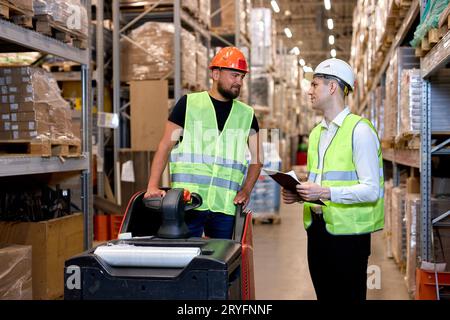 Young caucasian men workers doing stocktaking of products on shelves in warehouse together, using clipboard and forklift, having talk discussing, cont Stock Photo