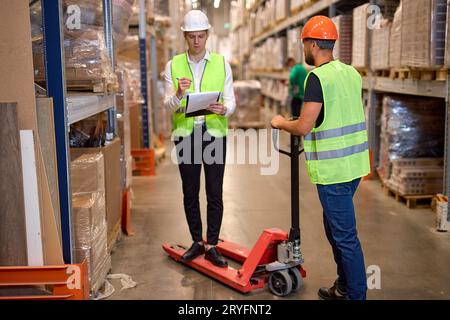 young warehouse workers in safety vasts and helmets talking together at work in large warehouse, discussing work, counting goods or cargo. wholesale, Stock Photo