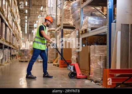 male staff worker in warehouse uses hand pallet stacker to transport goods, alone, dressed in working clothes and safety hard hat.Skilled warehouse em Stock Photo