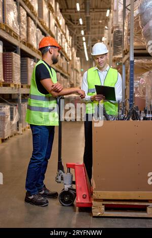 two men warehouse workers with hand pallet jack lift stand having talk, temwork. hand pallet truck and male mover. Workers in uniform with cargo cart. Stock Photo