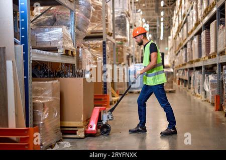 caucasian worker in a warehouse uses hand pallet stacker to transport pallets good cargo, hardworking confident male staff in green uniform vest at wo Stock Photo