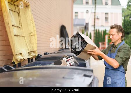 Throwing away unsorted trash employee man wearing apron put cardboard box with garbage plastic bag in it. Wrong unsorted garbage Stock Photo
