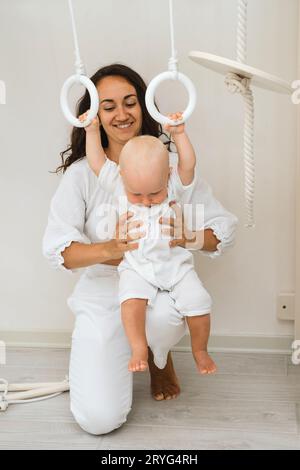 Baby is hanging on child Indoor Gym Playset on Wooden Gymnastics Rings. Kid swinging in playroom. Spending time on home sports complex active with Stock Photo