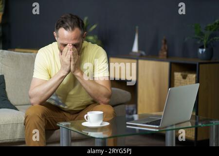 Middle age crisis, emotional stress mature man sits on sofa with hands covered his face next to coffee table with laptop and cup Stock Photo