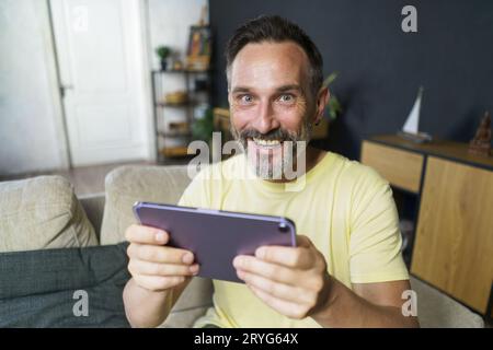 Excited playing games online middle aged man use digital tablet sitting on the sofa at home wearing casual. Mature freelancer ma Stock Photo