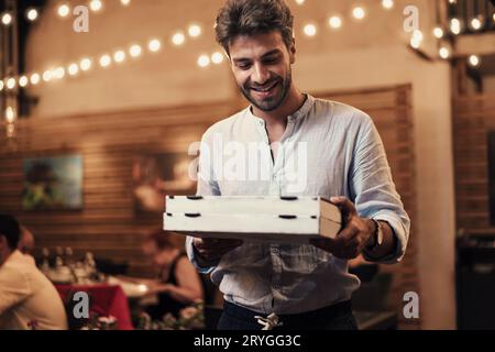 A man in his 30s exits a pizzeria at night, holding two takeaway pizza boxes. Ambient restaurant lights and tables illuminate the background. Stock Photo