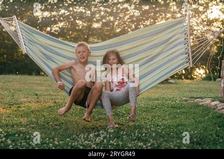 Happy children, boy and girl, brother and sister, friends laugh, swing in hammock on summer day in nature. Summer holidays in village. Games, entertai Stock Photo
