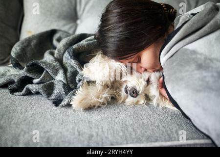 Young woman cuddling and kissing her dog while lying on couch at home Stock Photo