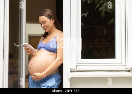 A pregnant woman is standing with a phone with a big belly in her house, using a mobile application. Space for text Stock Photo