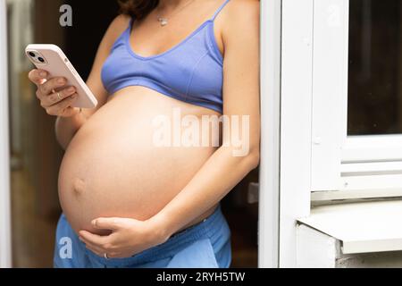 A pregnant woman is standing with a phone with a big belly in her house, using a mobile application. Space for text Stock Photo