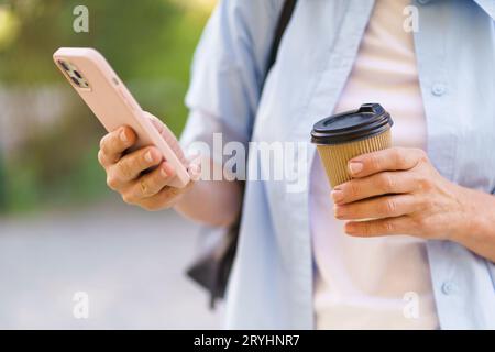 Close-up photo focuses on a woman's hands holding a mobile phone and a coffee paper cup. The image depicts modern lifestyle and Stock Photo