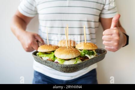 The guy holds a grill pan in his hands on which several juicy hamburgers lie and shows like. Delicious and fast food Stock Photo