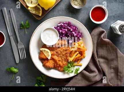 Schnitzel with potato fries, red cabbage salad and sauce on white plate over dark stone background. Top view, flat lay Stock Photo